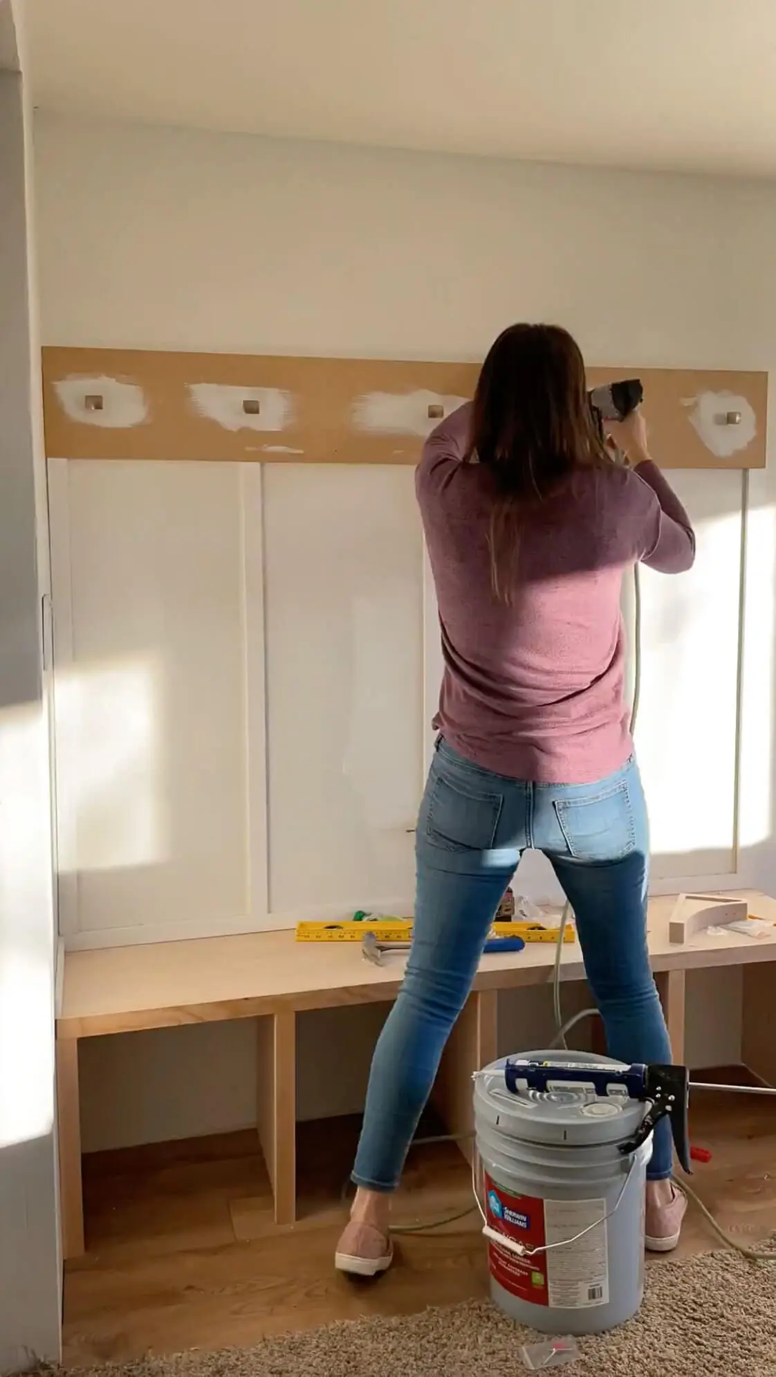 A woman securing a long wooden board to the wall above a built-in bench, using a nail gun. The space features a white paneled wall, wooden flooring, and a large bucket of construction adhesive nearby.